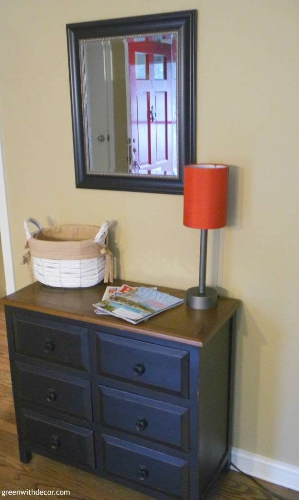 A small black dresser sits under a mirror with a red lamp.