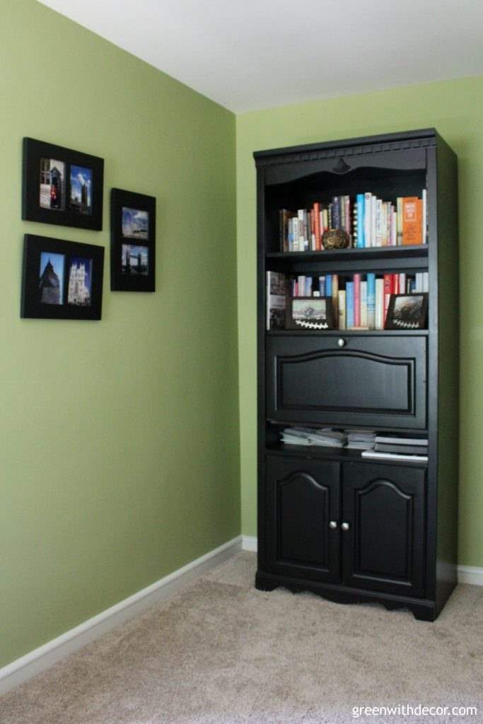 A black bookshelf full of books. Walls are Ryegrass green with black picture frames on the wall.