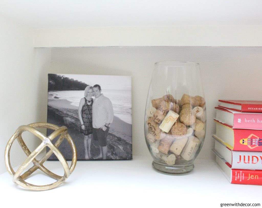 A white painted bookshelf with a gold decorative sphere, a black and white picture, corks and colorful books.