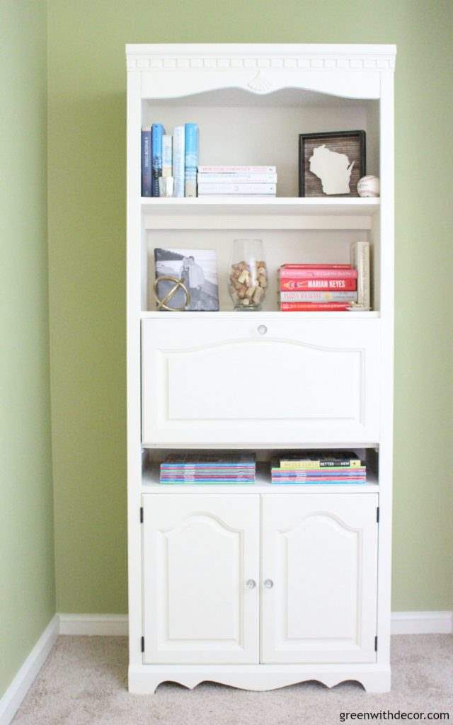 A white clay painted bookshelf with colorful books, aWisconsin sign and a gold decorative sphere. Wall color is Ryegrass.