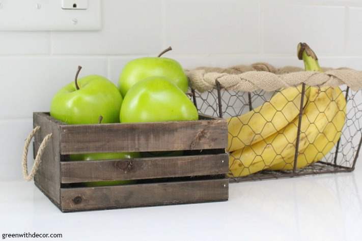 Wooden crate with green apples and wire basket with bananas on white quartz counter with white subway tile background