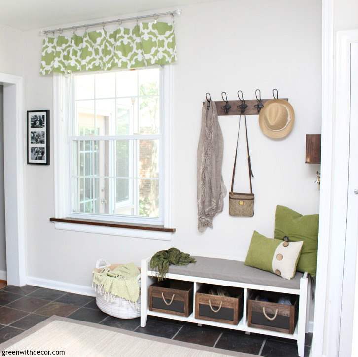 A large window with green and white valance next to a bench with storage and hooks on the wall