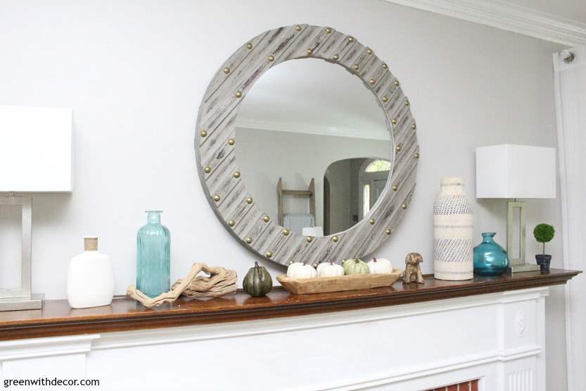 A round grey mirror hangs above a fireplace with coastal fall decor displayed