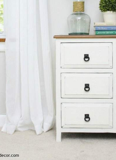 A short white dresser with a wood top and black hardware. Agreeable Gray walls with white curtains, white trim and tan carpet.