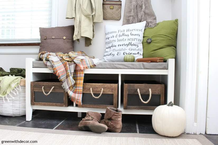 A white bench with wood crates for shoe storage and throw pillows on top of the bench.