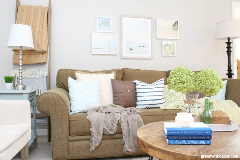 A coastal casual living room with Agreeable Gray walls, a dark brown couch, green and blue throw pillows, a beachy gallery wall and a round rustic wood coffee table.