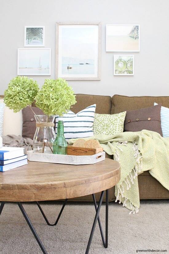 A coastal casual living room with Agreeable Gray walls, a dark brown couch, green and blue throw pillows, a beachy gallery wall and a round rustic wood coffee table with dried green hydrangeas.