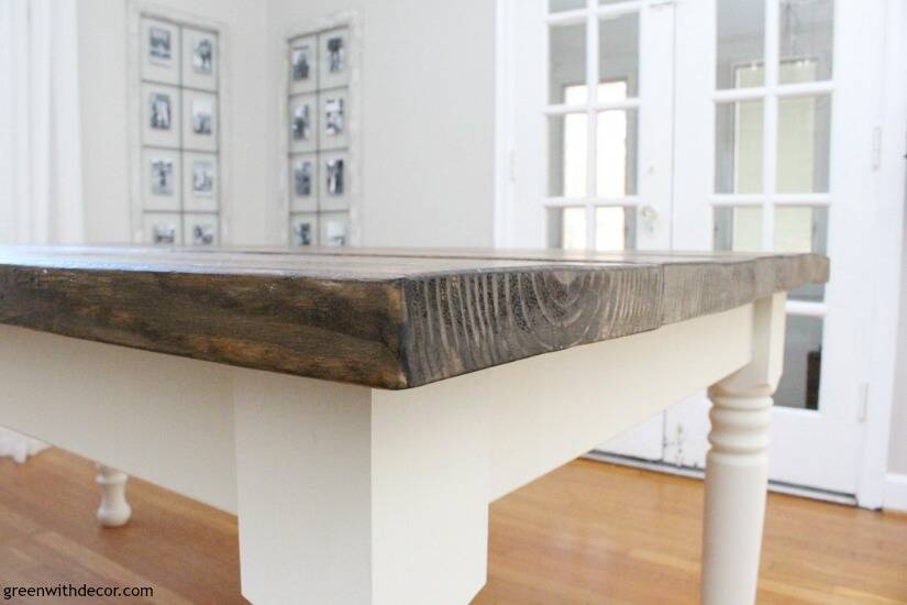 The corner of dark stained wood farmhouse table with white spindle legs, sitting on a hardwood floor with white French doors behind it.