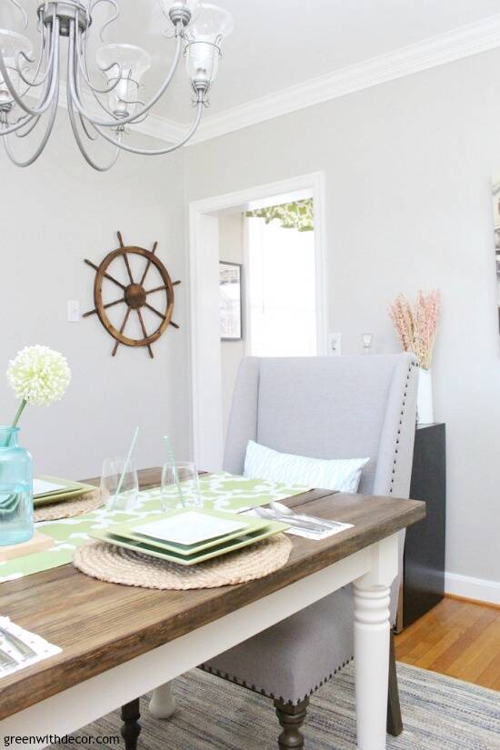 A dark wood farmhouse dining table with white legs, a gray fabric chair at the end, gray walls with a wooden ship wheel.