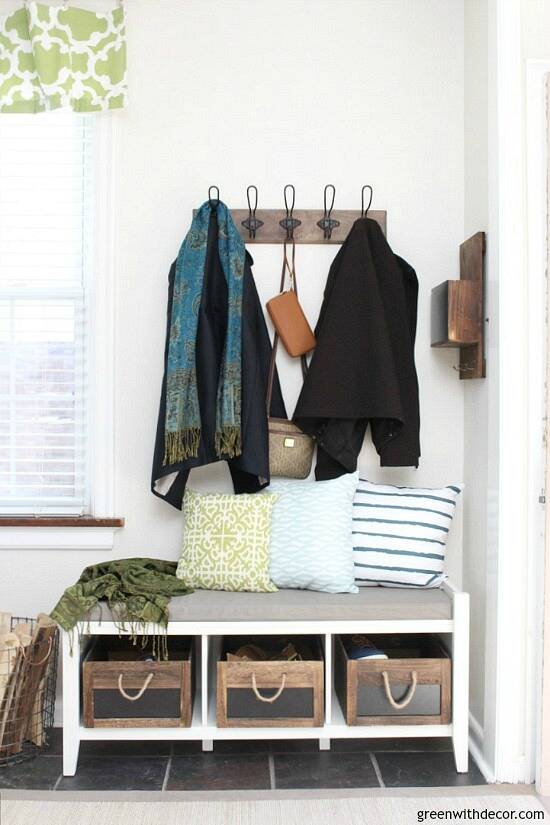 A foyer entry with a white bench, green and blue pillows and coats hung on the wall