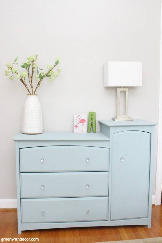 A blue three-drawer dresser combo with a white vase, flamingo sign, green clothespin and silver lamp on top. The walls are Agreeable Gray.