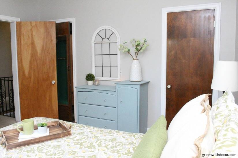 A neutral, blue and green bedroom with gray walls, a blue dresser and old white window on the wall.