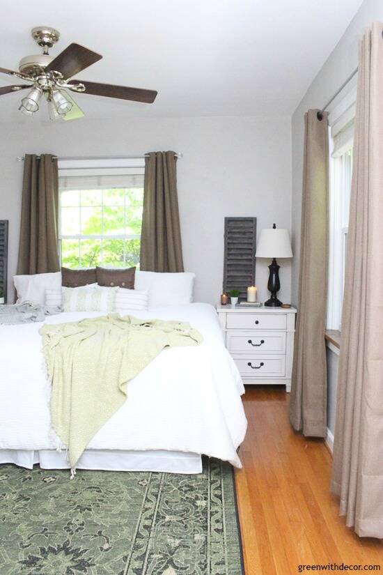 A neutral rustic bedroom with gray walls, white bedding, a green rug and brown curtains